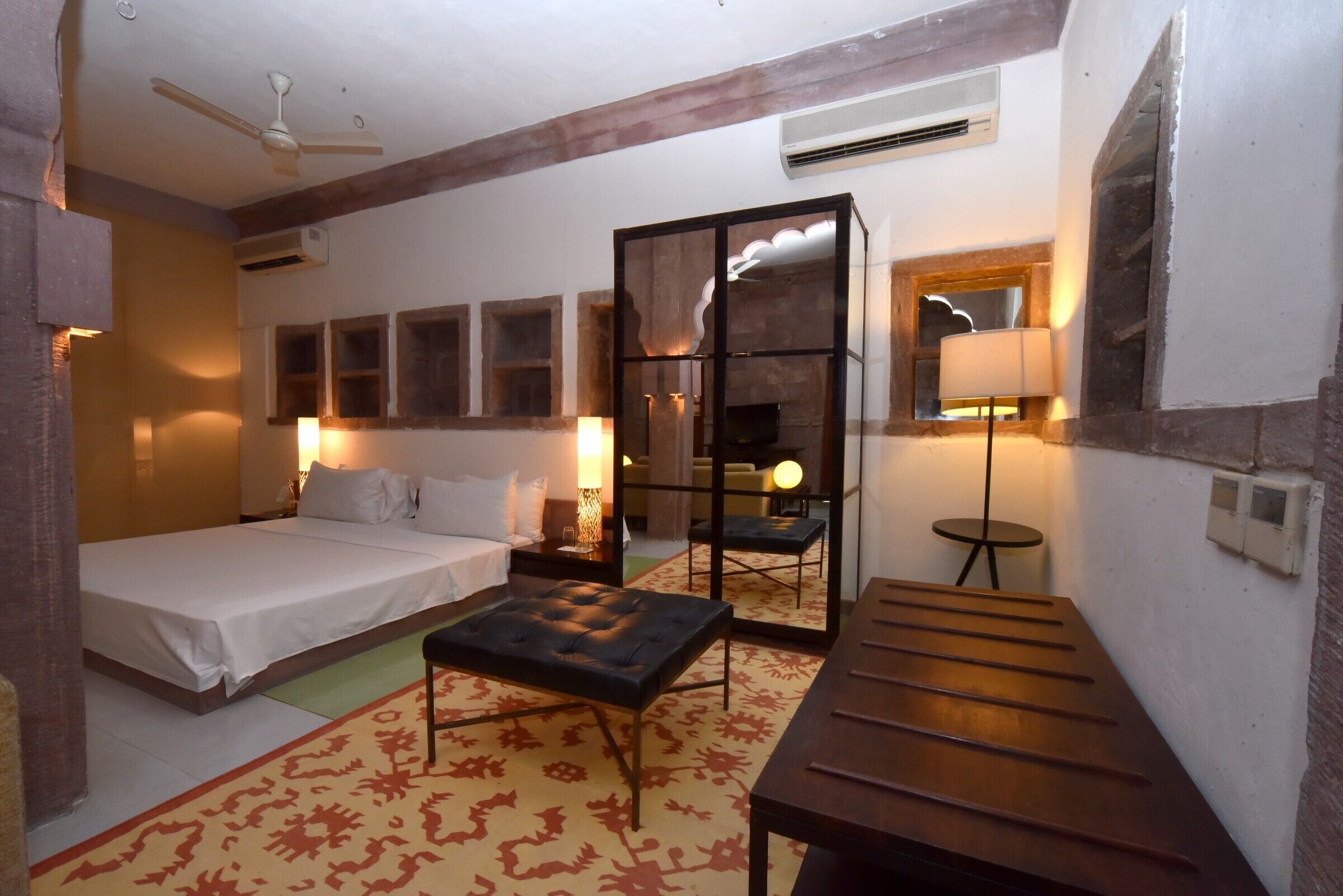 Una habitación de hotel con una cama, un banco de madera, una mesita, un espejo y una lámpara.