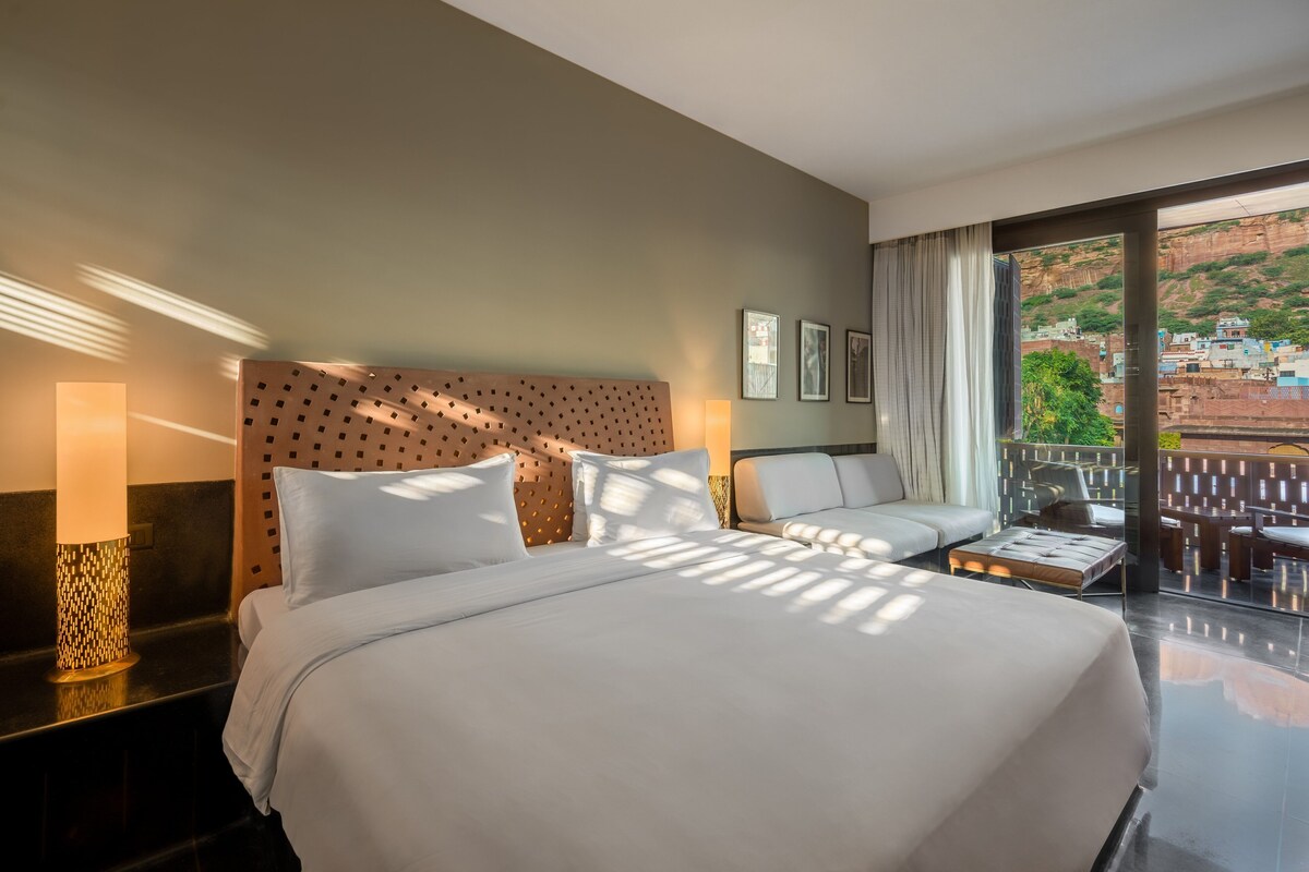 A hotel room with a large bed, a sofa, a balcony with a view of the city, and a patterned headboard.