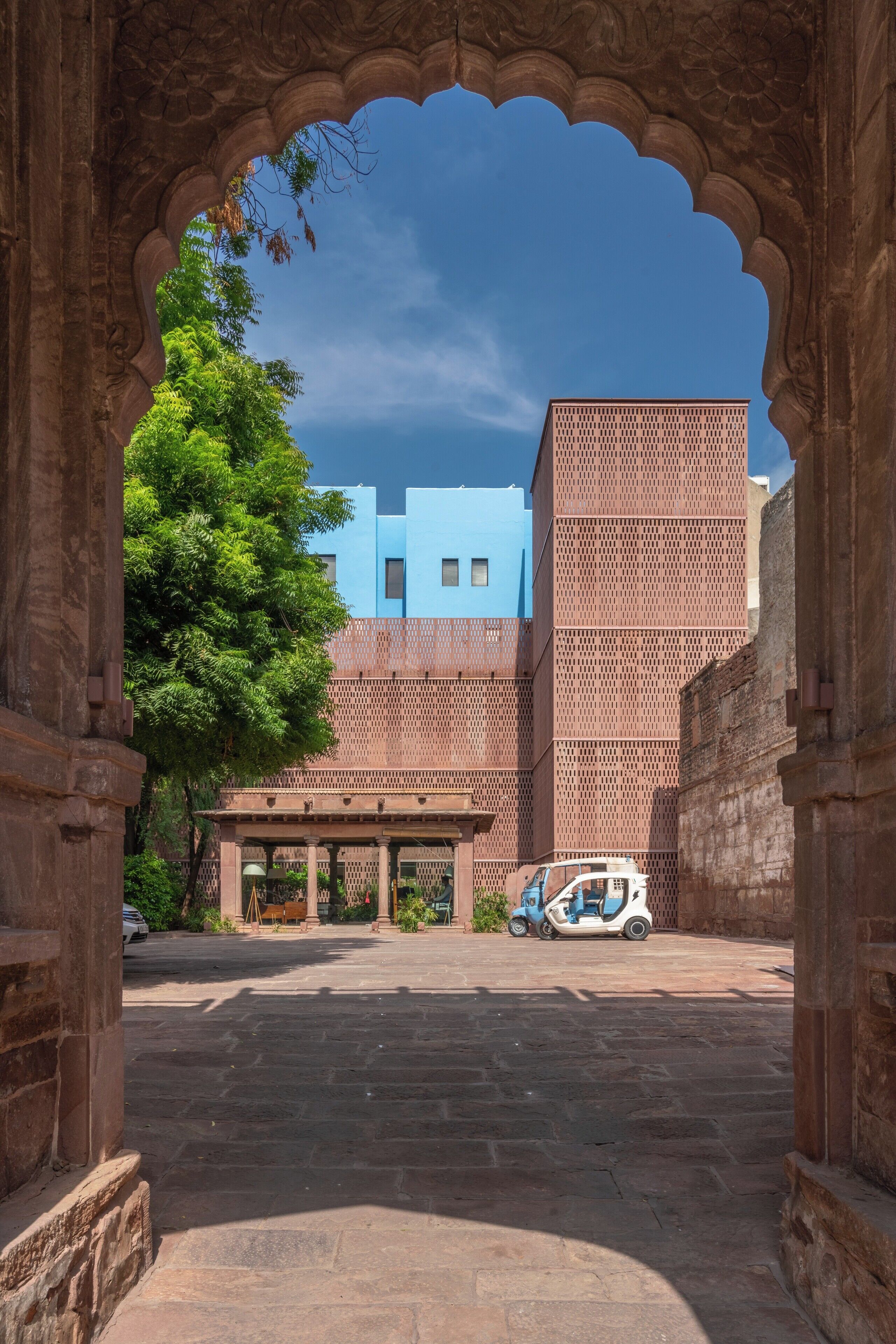 An arched doorway leading to a courtyard with a modern building in the background, a tree, and a parked scooter.