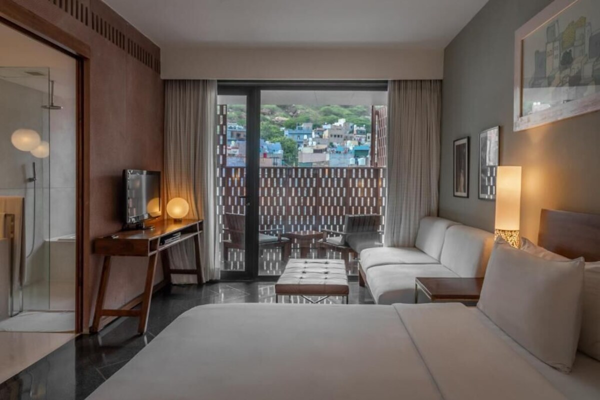 A hotel room with a bed, a desk, a sofa, and a balcony with a city view.