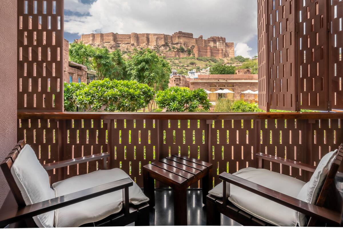 A balcony with two wooden tables and chairs, a view of a historic fort, and lush greenery.