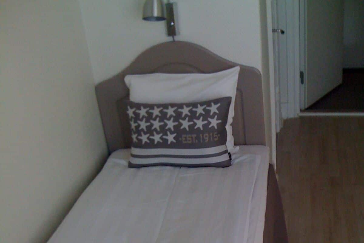 Une petite chambre avec un lit simple, une tête de lit, une lampe de chevet et une porte donnant sur une autre pièce.