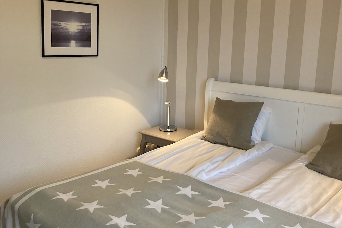 Un lit bien fait, recouvert d’une couverture à motifs étoilés, une lampe de chevet et un tableau encadré au mur.