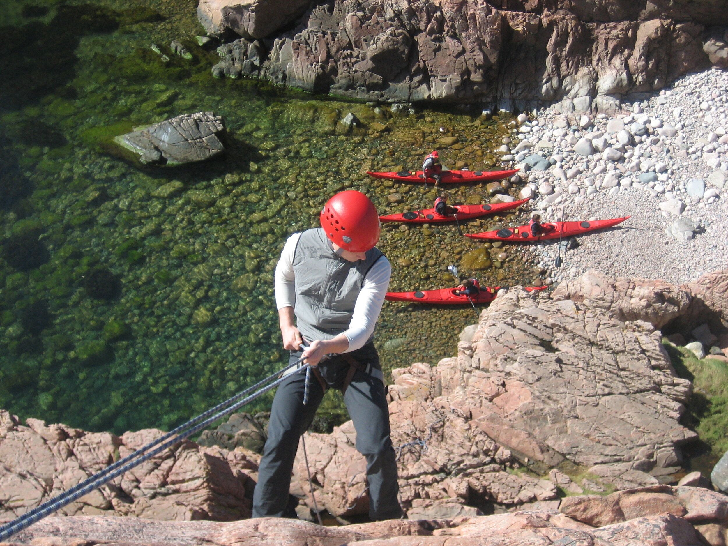 Un kayakista preparando una cuerda en una orilla rocosa con agua cristalina y otros kayakistas al fondo.
