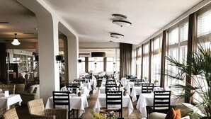 Un hall d’hôtel spacieux, doté de hauts plafonds, de grandes fenêtres et d’un espace salon central avec une table basse et des fauteuils en osier.