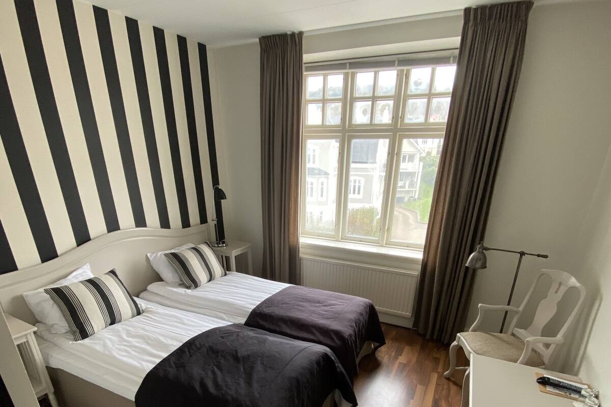 Une chambre d’hôtel avec deux lits, un bureau et une chaise. Il y a une fenêtre avec des rideaux et une vue sur les bâtiments environnants.