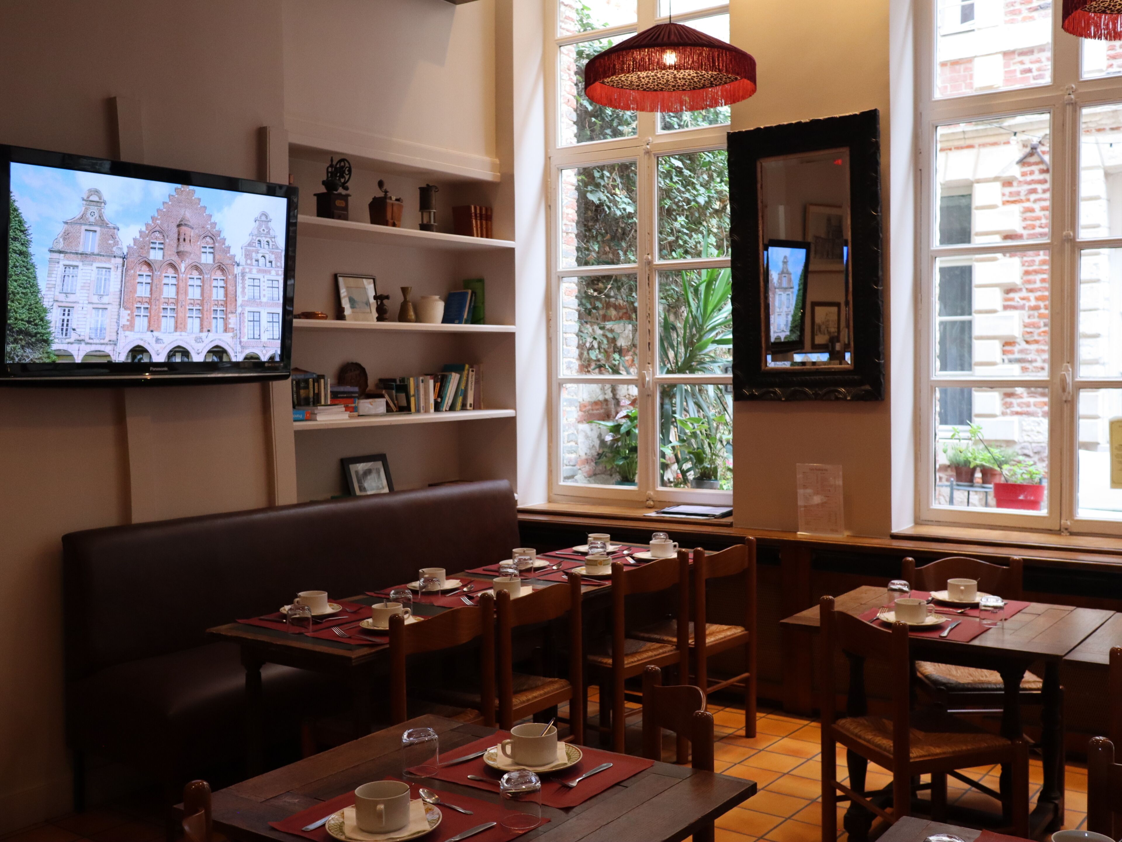 A dining area with tables set for a meal, a television mounted on the wall, and a window with a view of a building.