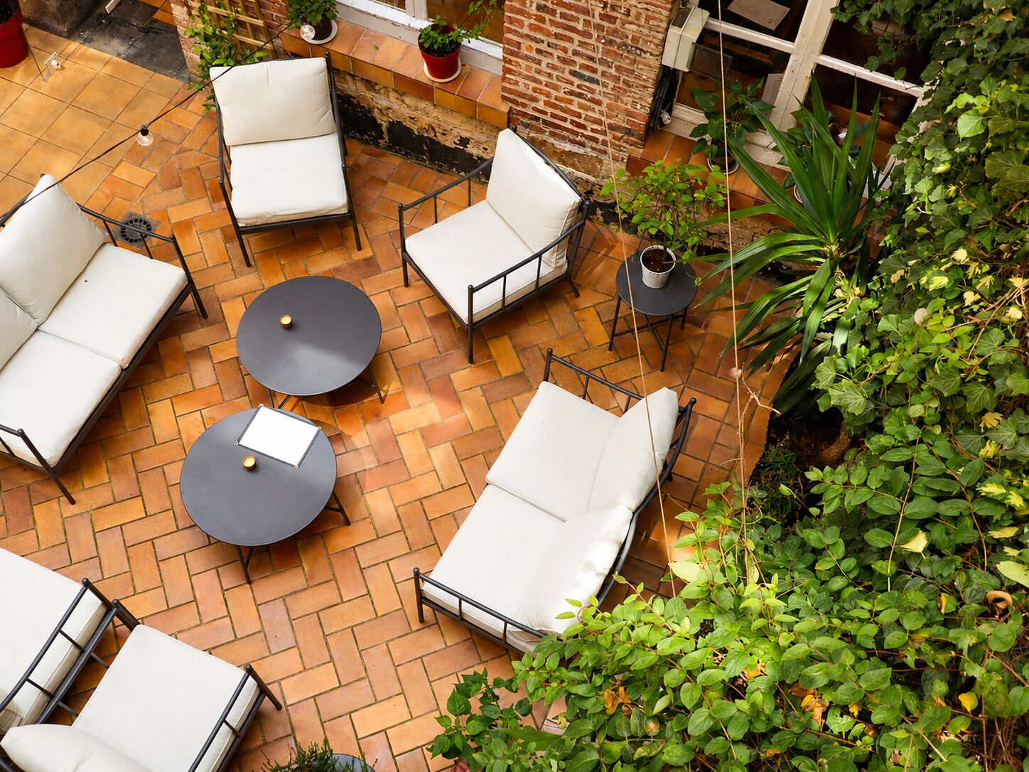 An outdoor patio with multiple white cushioned seating areas and round tables, surrounded by potted plants and a brick wall.