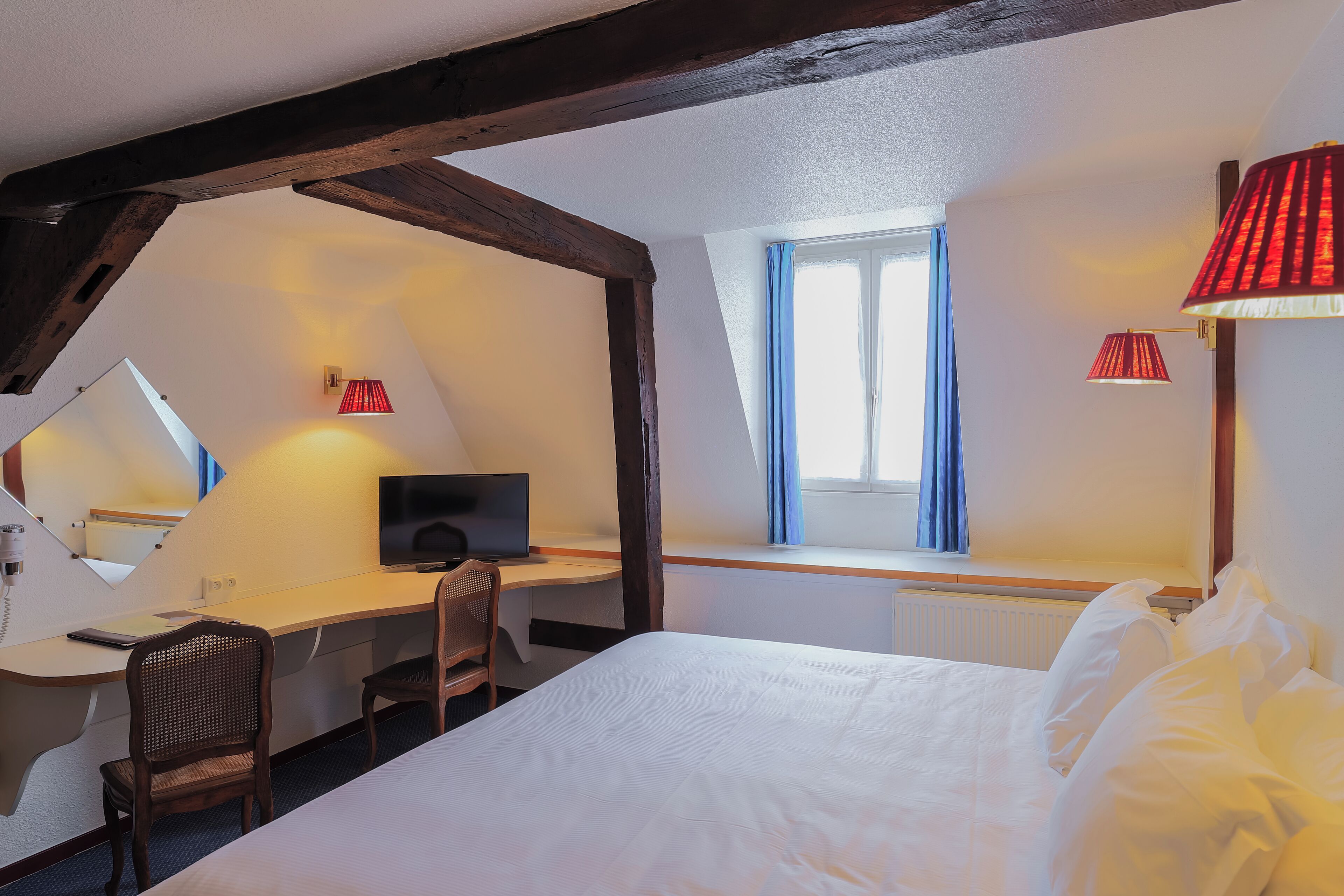 Habitación de hotel con cama, escritorio, silla, televisor y ventana con cortinas.