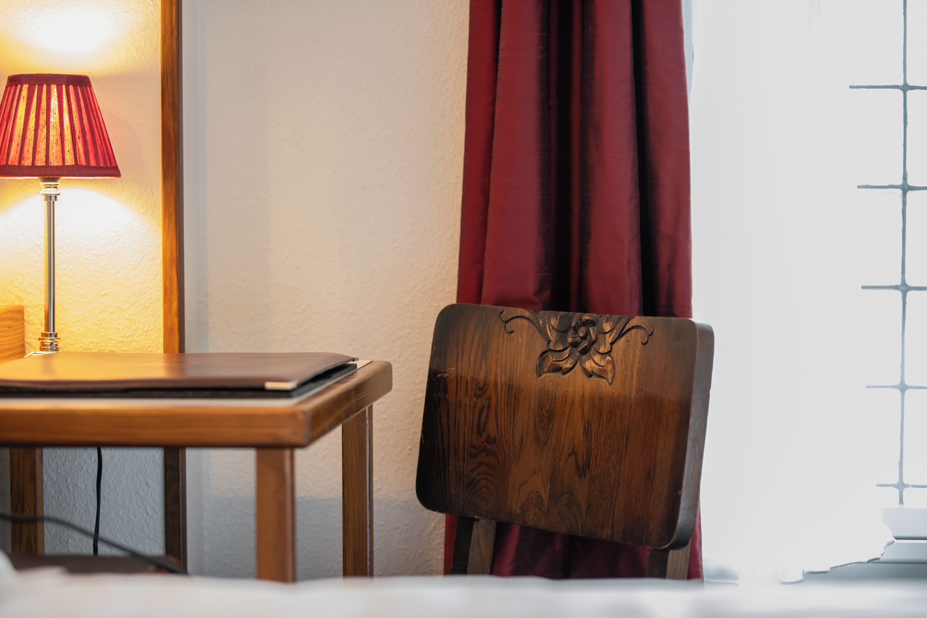 Uma cadeira de madeira com encosto decorativo, uma mesa com abajur e uma janela com padrão quadriculado.