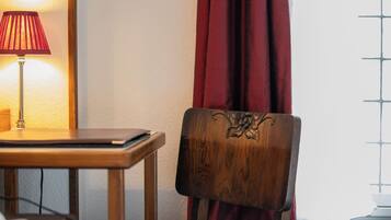 Uma cadeira de madeira com encosto decorativo, uma mesa com abajur e uma janela com padrão quadriculado.