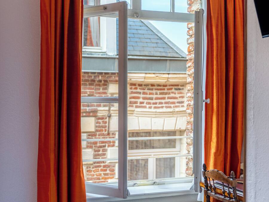 A hotel room with a bed, a rolled white towel, orange curtains, and a view of a brick building through a window.
