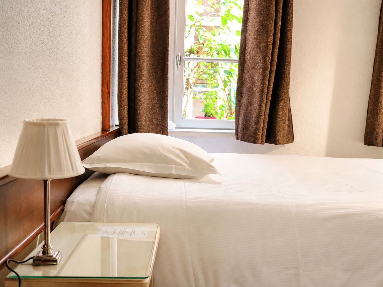 A hotel room with a bed, a nightstand, a lamp, and a window with curtains.