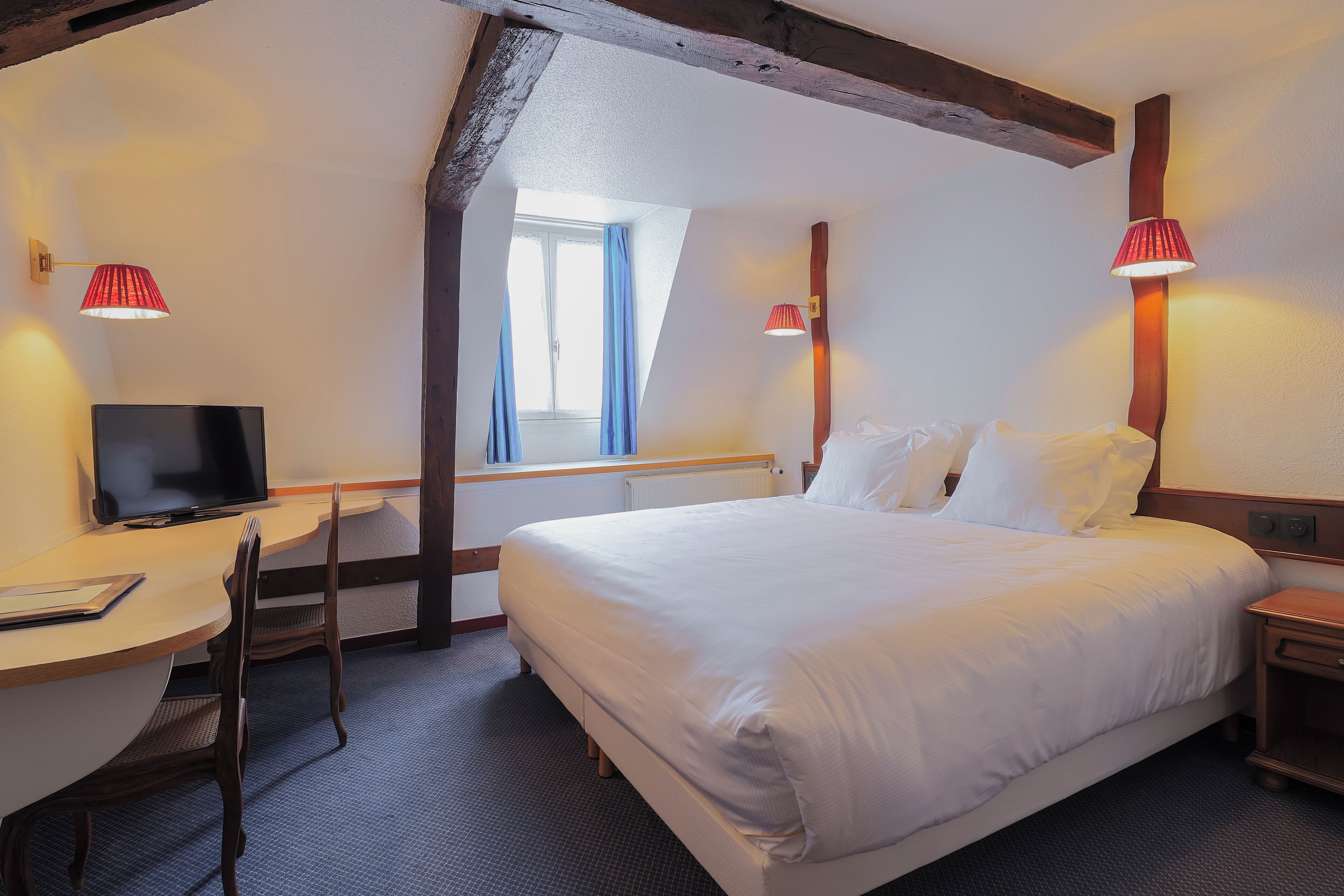 A hotel room with a bed, a desk with a chair, a television, and two wall-mounted lamps.