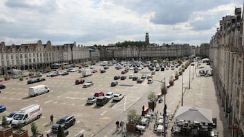 A large parking lot with multiple cars and a few pedestrians. There are buildings with classical architecture in the background.