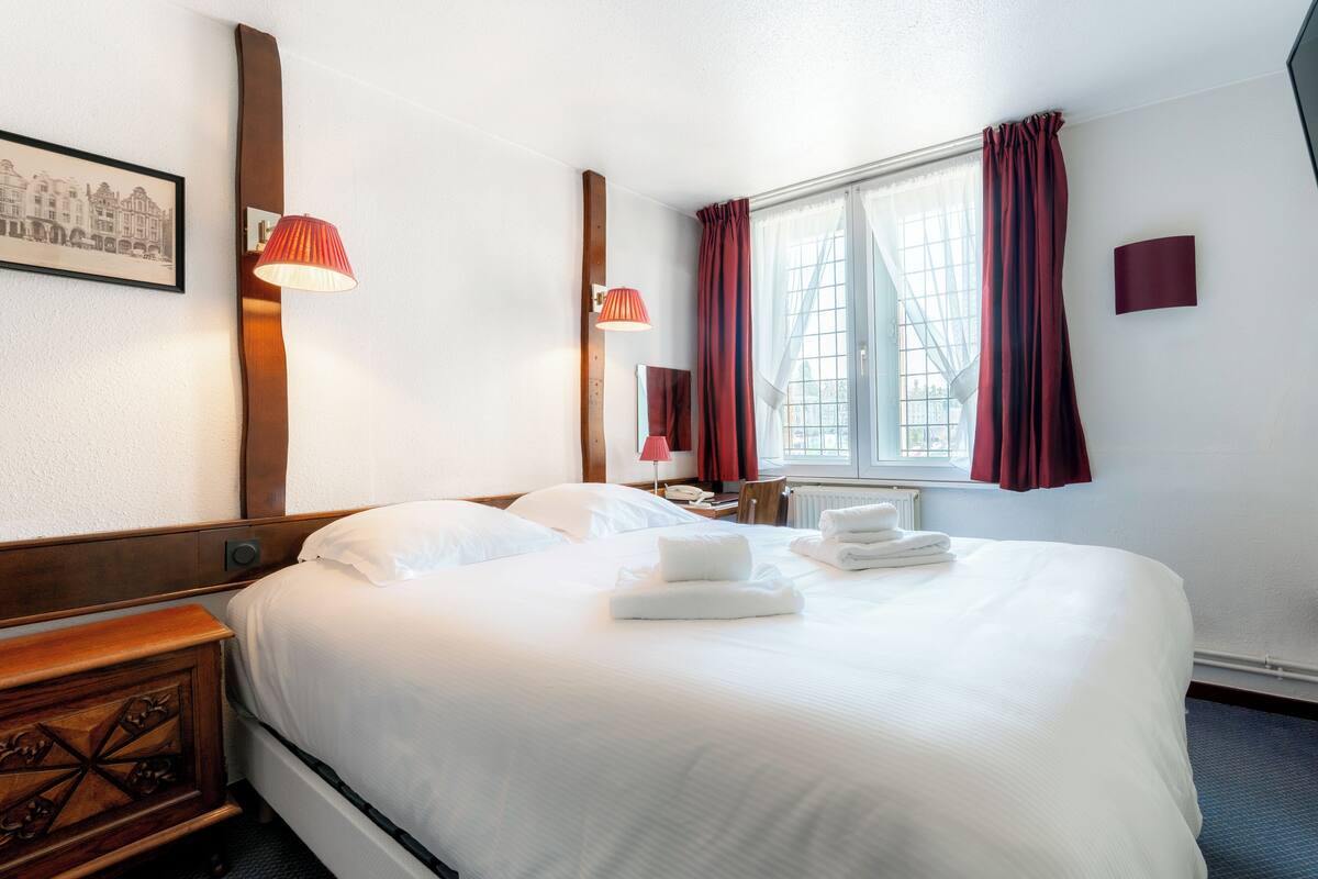 Habitación de hotel con una cama grande, una mesita de noche de madera, una mesita auxiliar con lámpara, una ventana con cortinas y un cuadro en la pared.