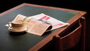 Een tafel met een kop en schotel, een krant en een raam met uitzicht op groen.