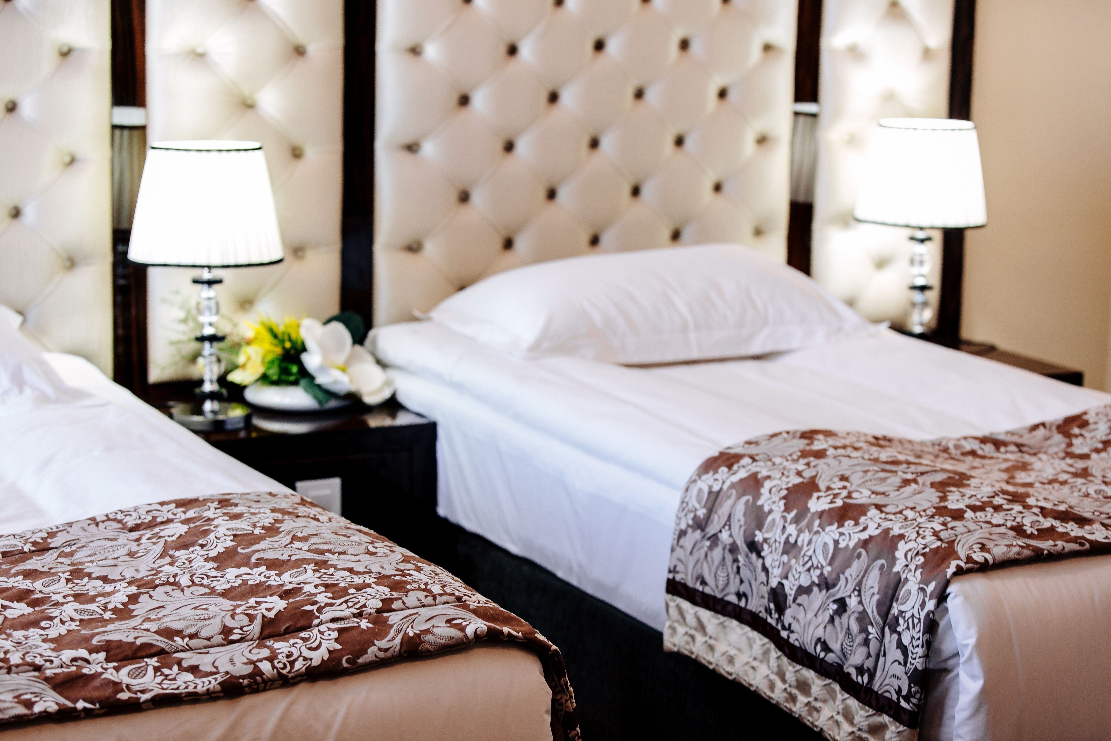 A hotel room with two beds, each with a white bedspread and a brown patterned throw. There are two bedside tables with lamps, and a decorative vase with flowers on one table.