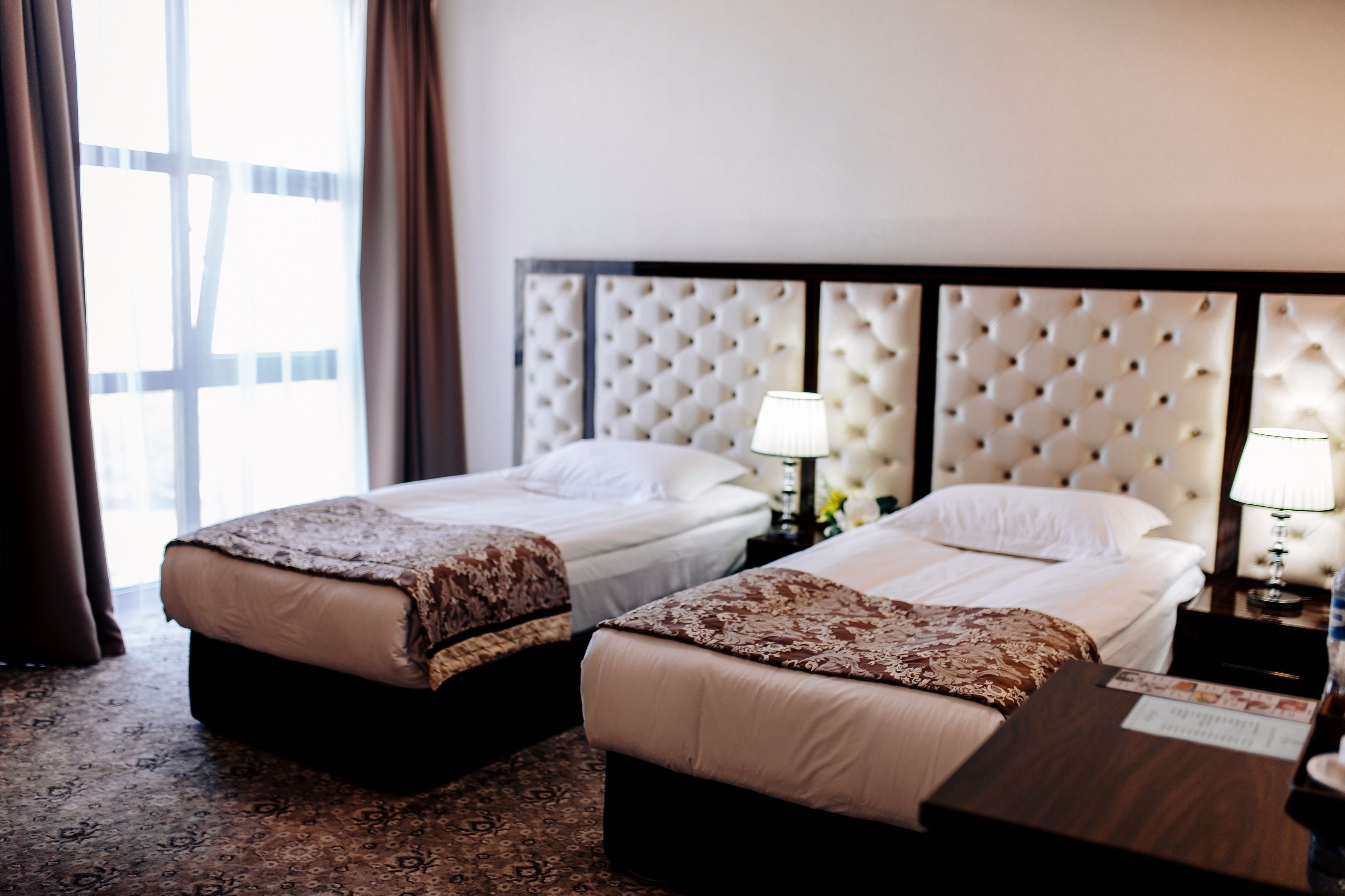 A hotel room with two beds, a large window, and a tufted headboard.