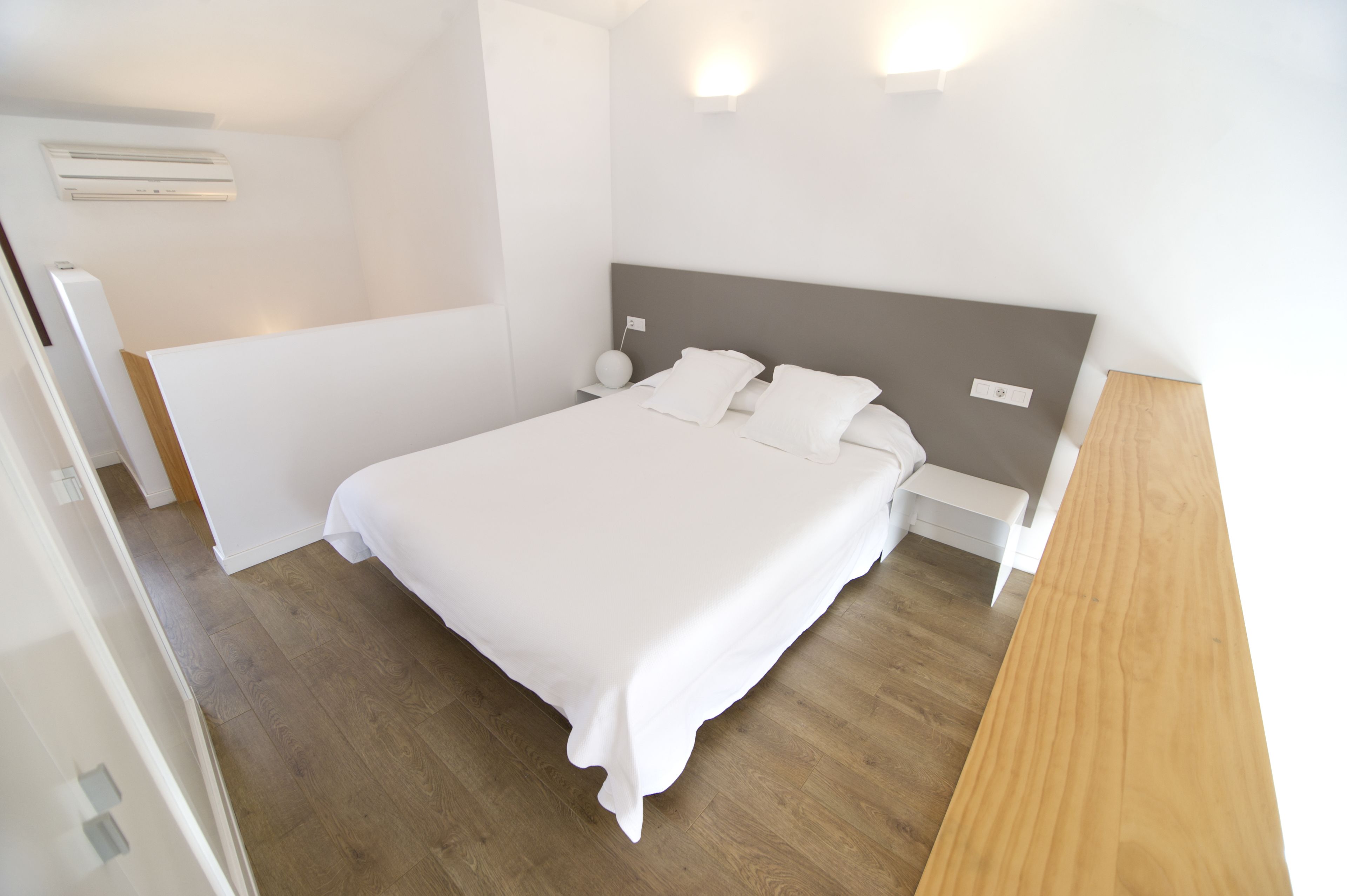 A neatly made bed in a hotel room with a wooden floor, a bedside table, and a wall-mounted light fixture.