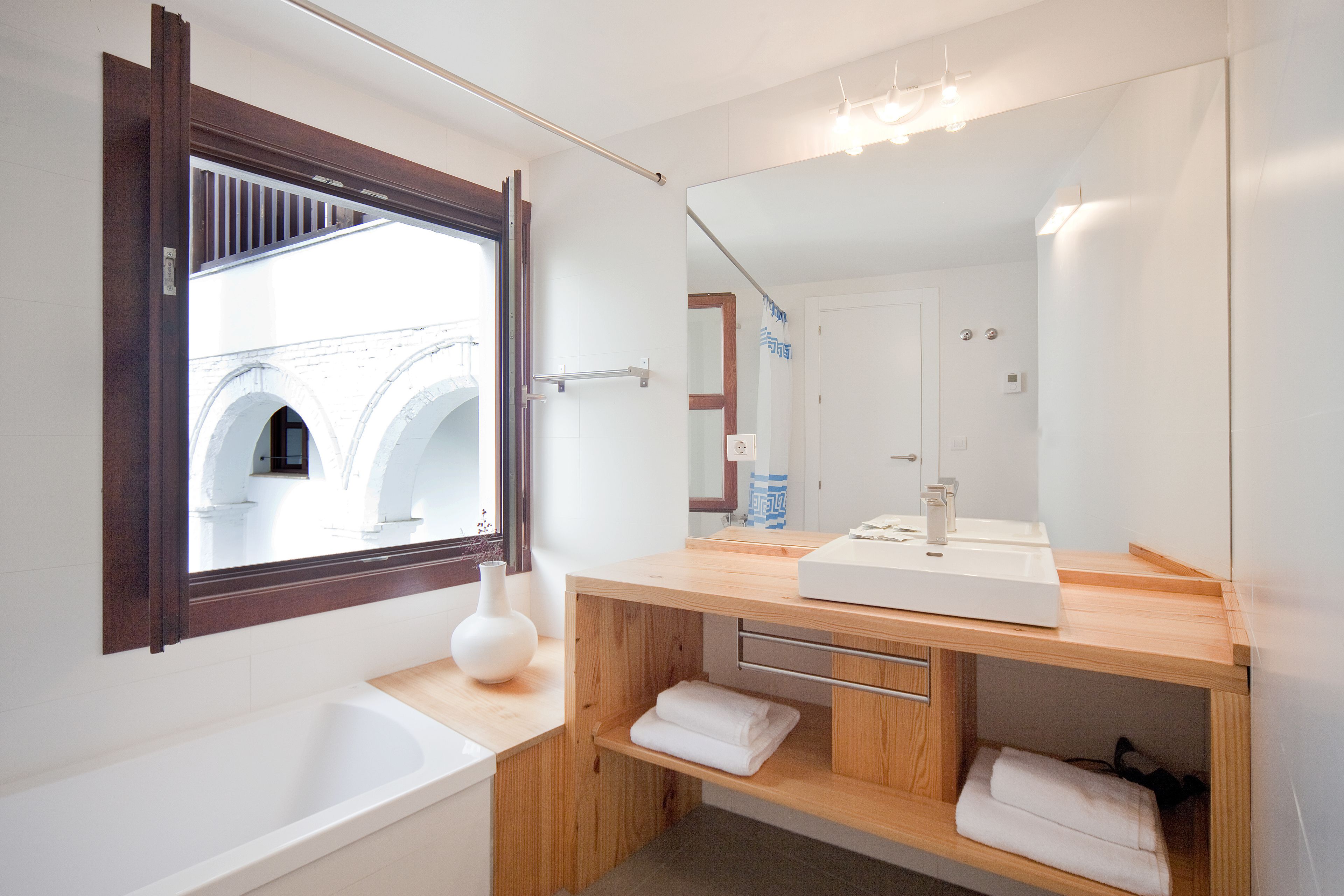 A modern bathroom with a wooden vanity, a large mirror, a white bathtub, and a view of an arched window.