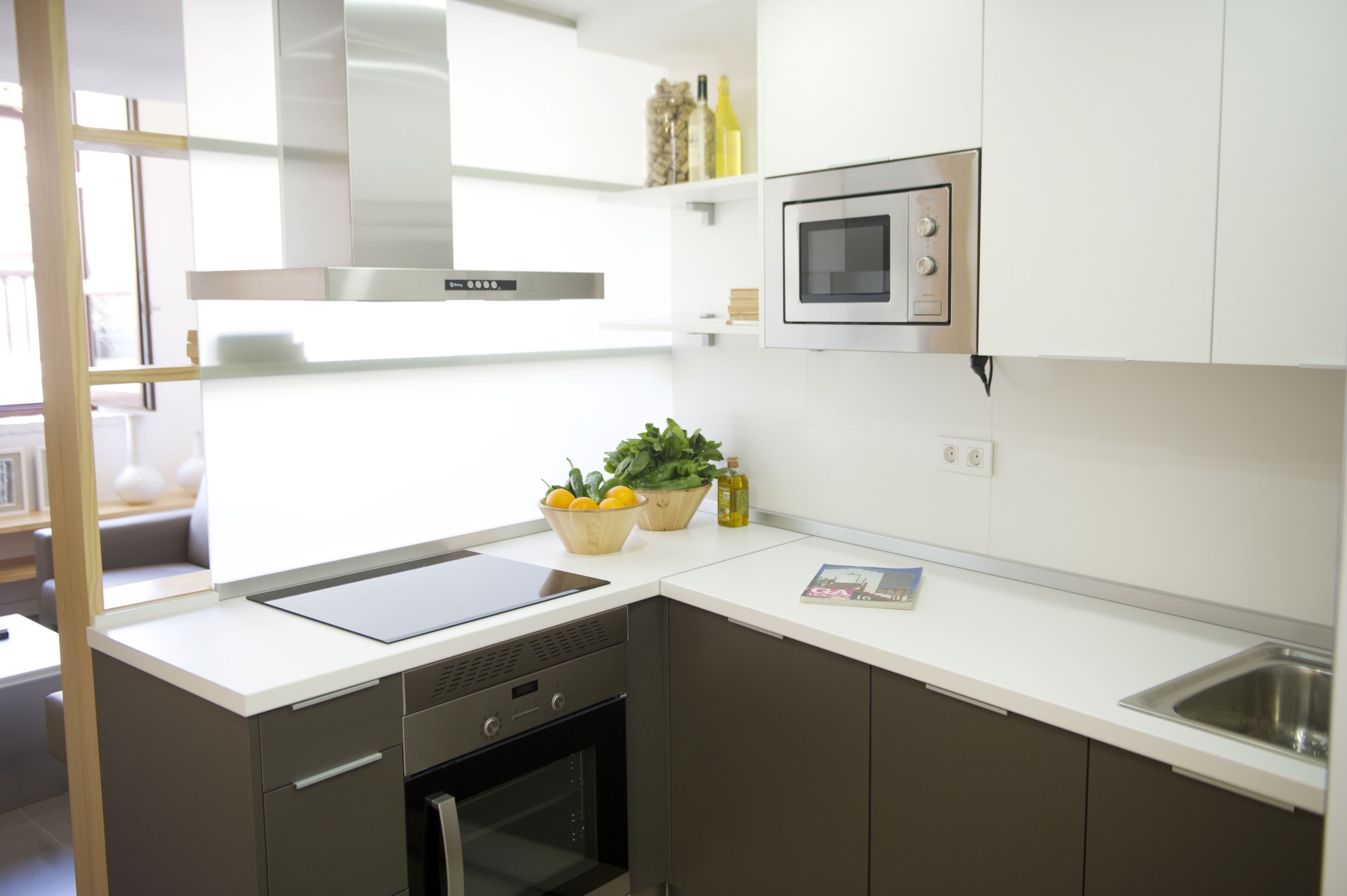A modern kitchen with a built-in oven, microwave, and stainless steel range hood. There is a small island with a sink and a bowl of lemons and a plant.