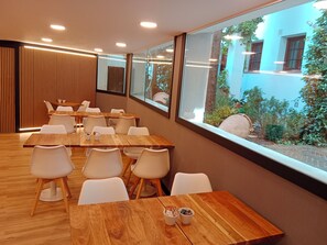 Un comedor moderno con mesas de madera y sillas blancas, ventanas amplias que dan a vistas de vegetación y un edificio exterior.