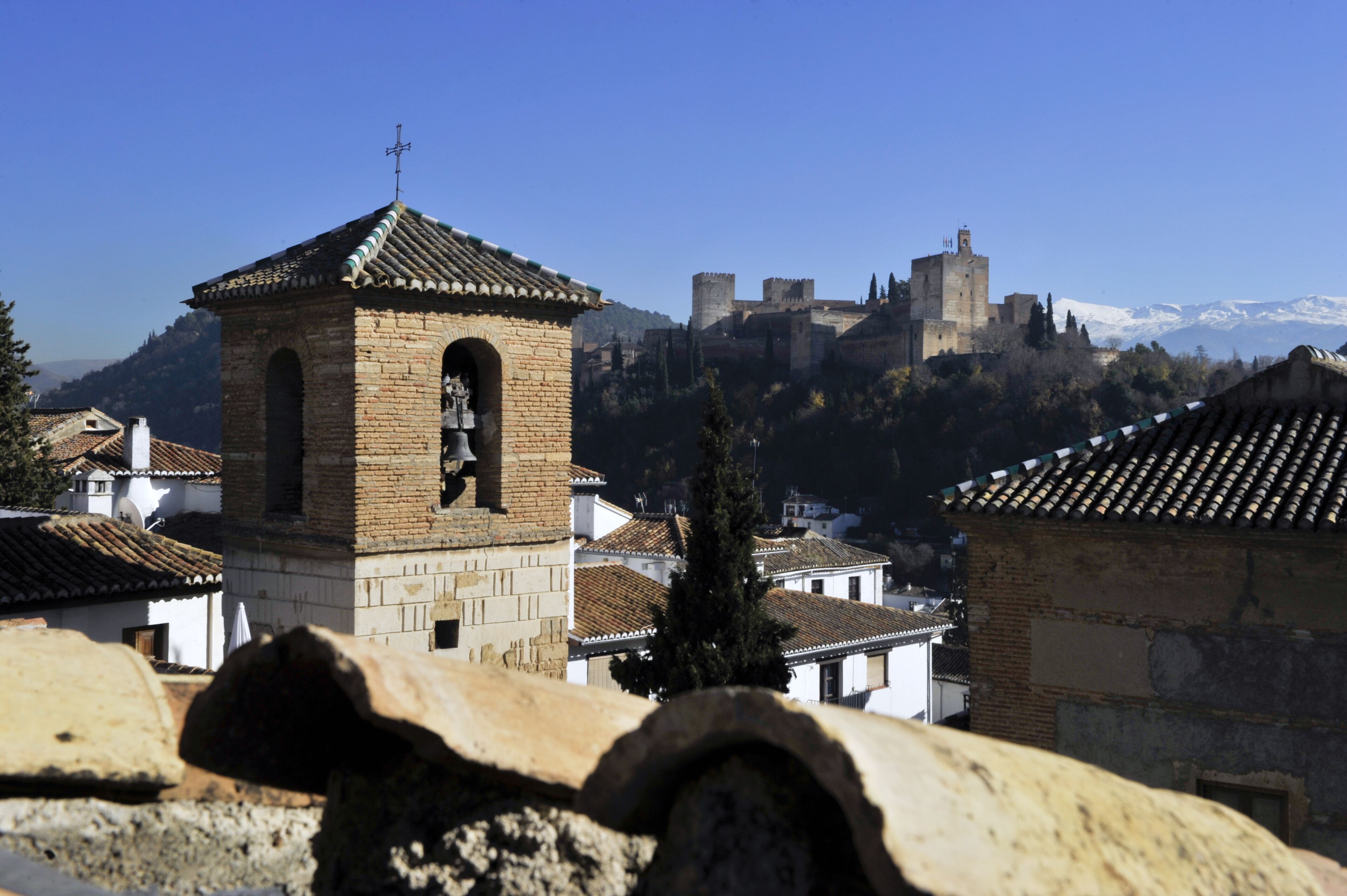 Una torre de piedra histórica con una campana, un castillo al fondo y un paisaje montañoso.