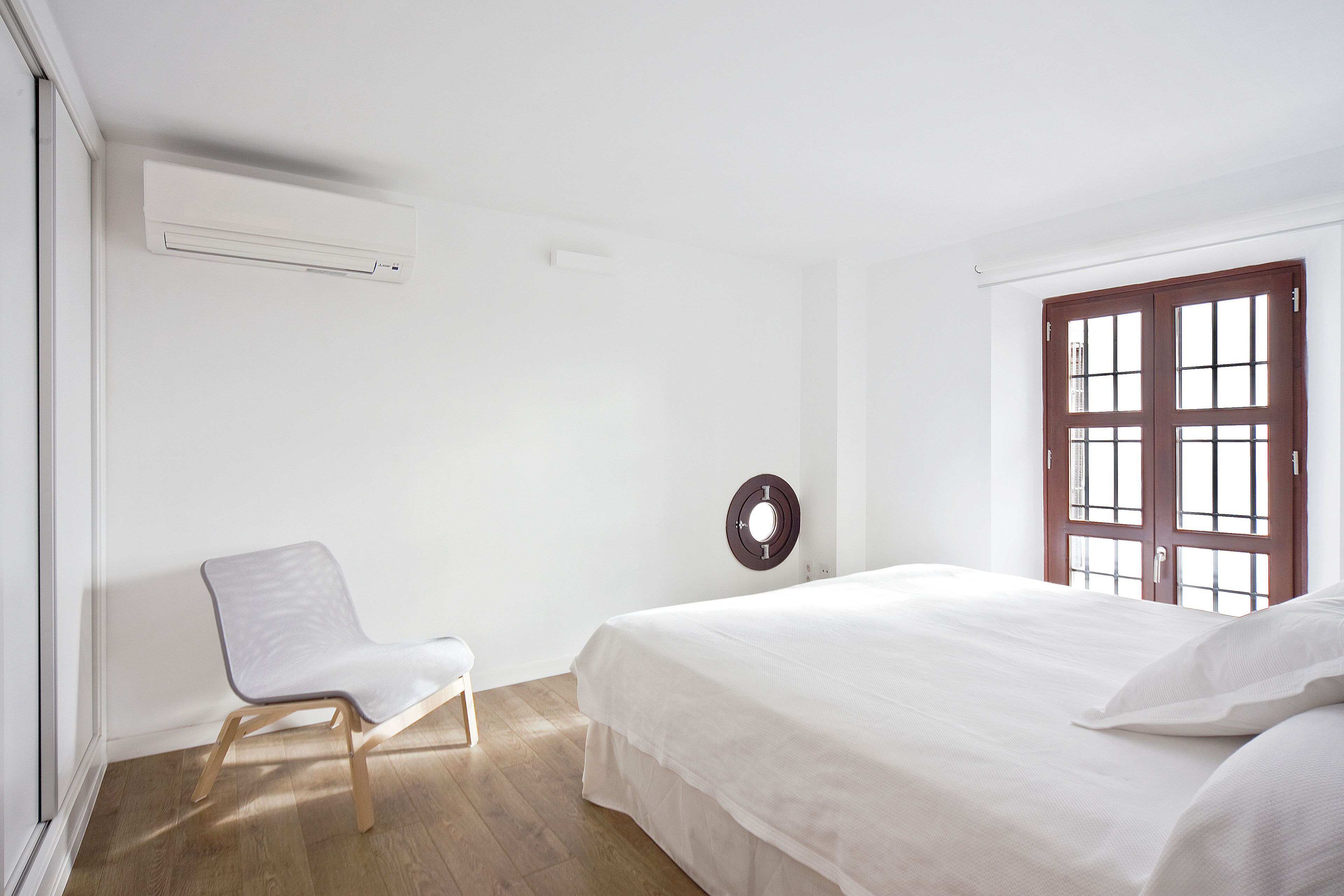 Une chambre à coucher minimaliste, comprenant un lit, une chaise, un climatiseur fixé au mur et une fenêtre avec des volets en bois.