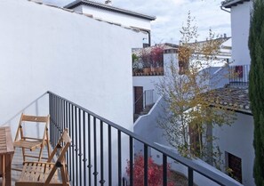 Un balcón con sillas y mesa de madera, paredes blancas y un árbol de hojas rojas.