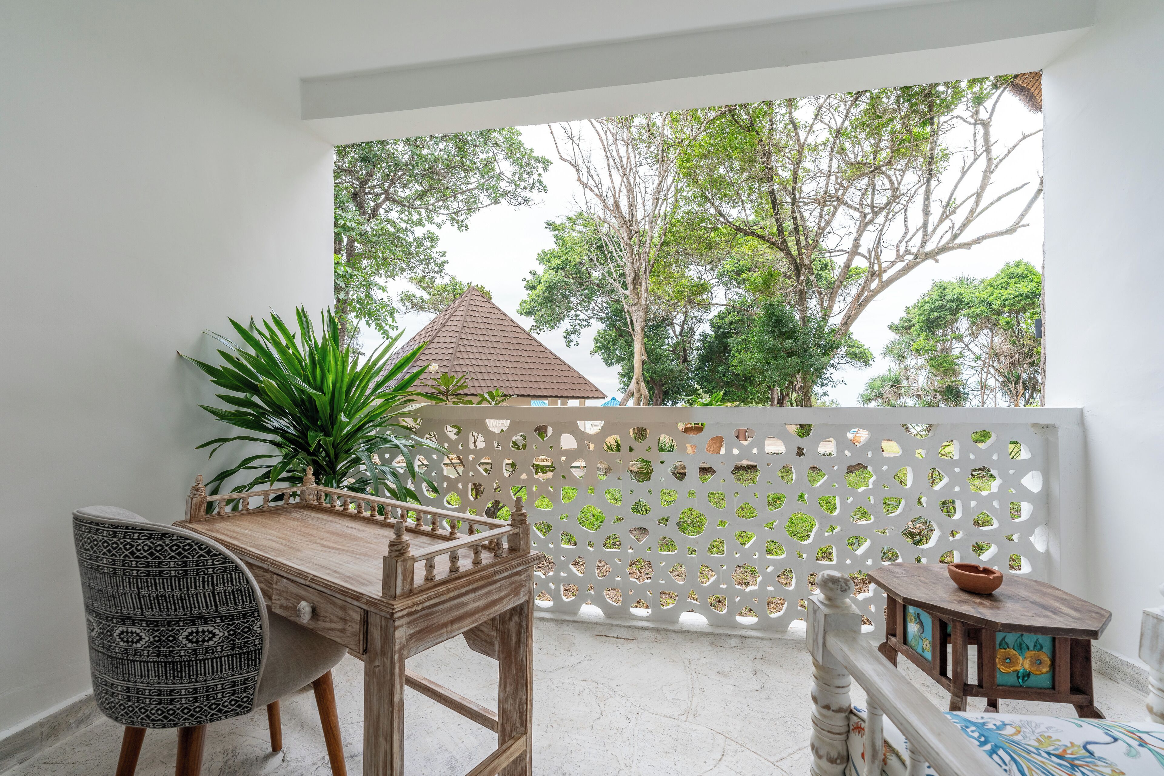 A balcony with a wooden table and chairs, a decorative fence, and a view of trees and a building.