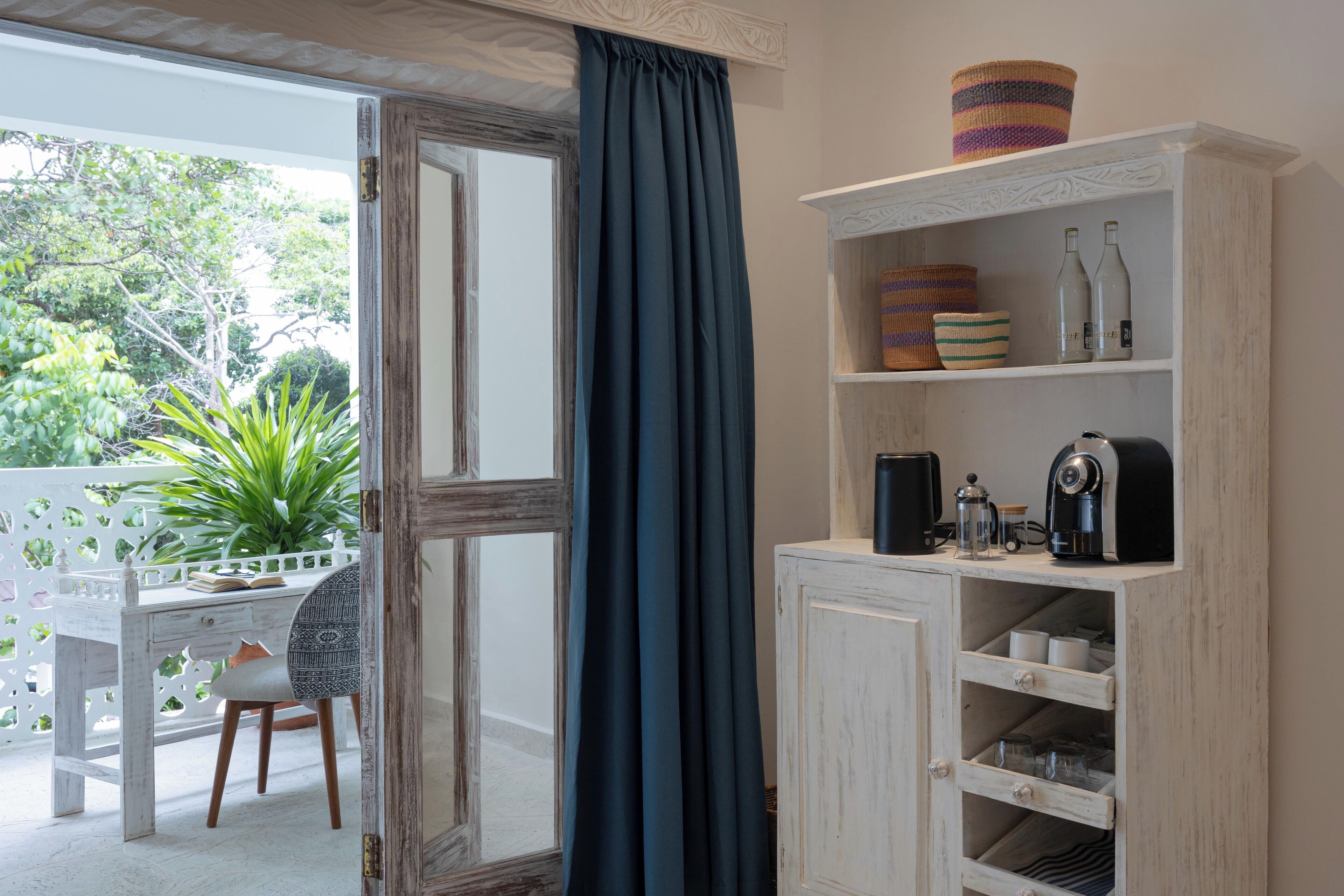 Una habitación con un escritorio de madera blanca, una silla, una estantería con diversos objetos y vistas al exterior con vegetación.