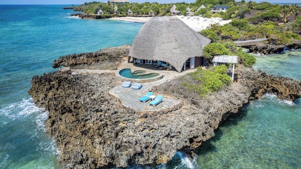 Un resort isleño tropical con un edificio de techo de paja, aguas azul cristalino y una costa rocosa.