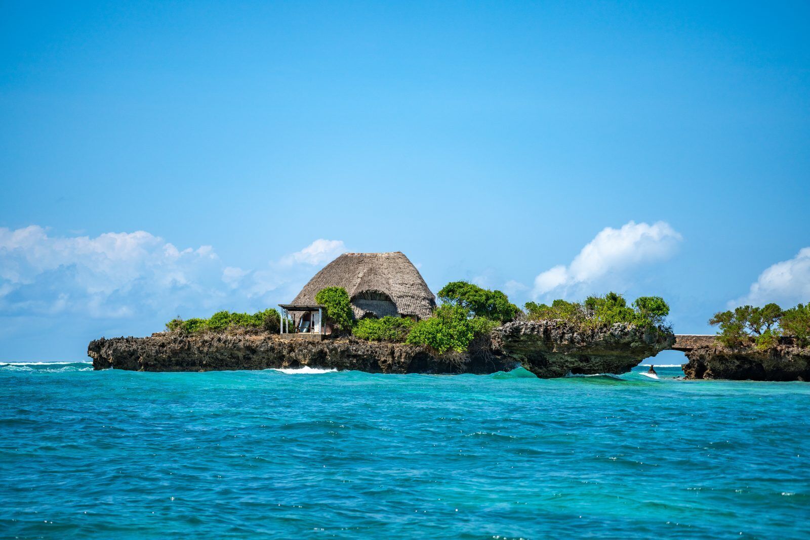 A thatched-roof hut with a small porch, surrounded by lush greenery on a rocky outcrop in clear blue waters.