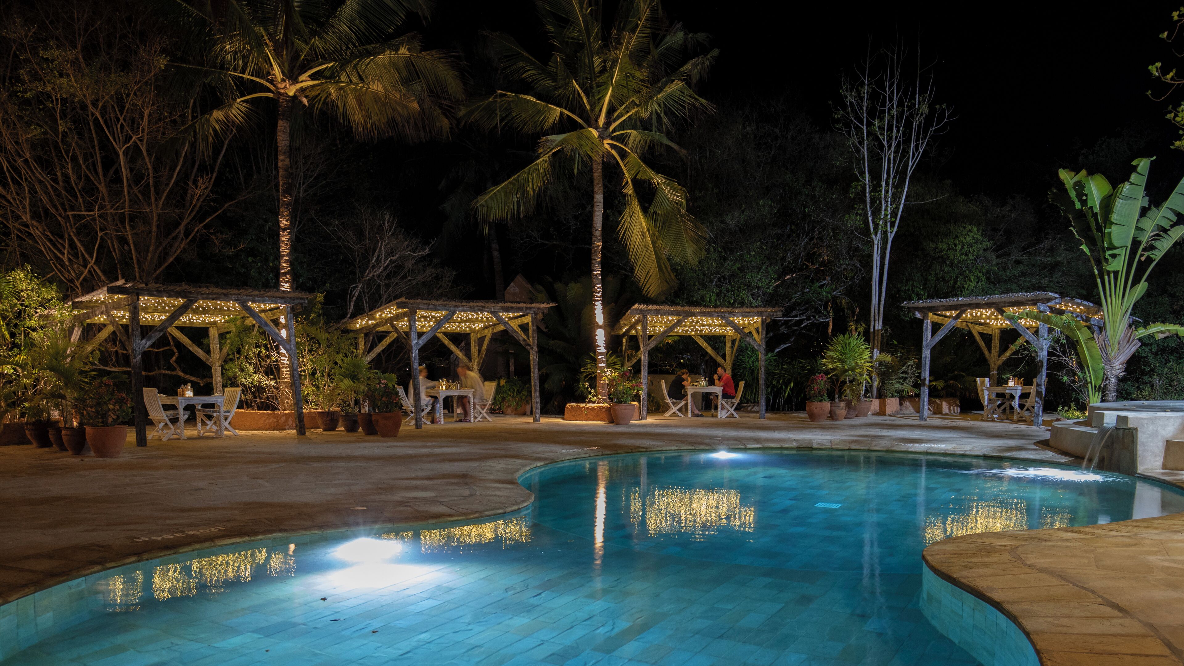 A nighttime pool area with a curved pool, wooden gazebos, and potted plants.