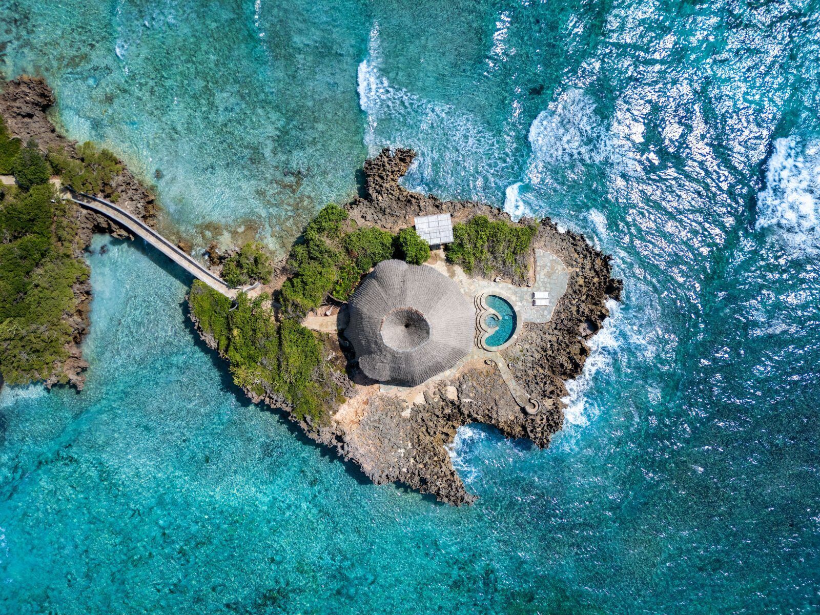 An aerial view of a secluded island with a circular stone structure, a small pool, and a pathway leading to a building.