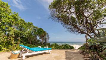 A sun lounger with a blue cushion and a woven bag on a stone patio overlooking the ocean.