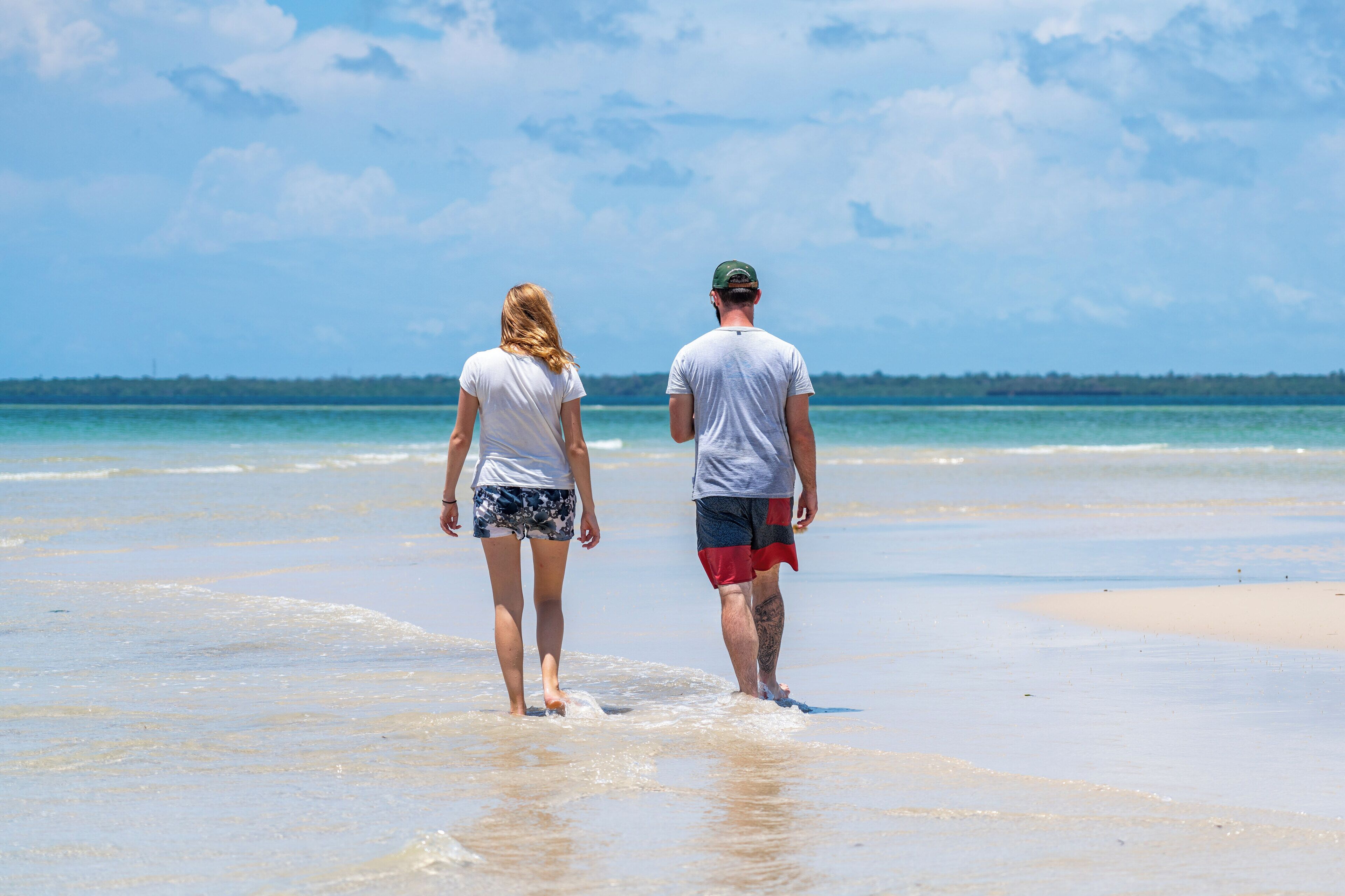 Two people walking on a sandy beach with a clear sky and calm sea in the background.