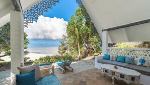 Un espacio de estar al aire libre con techo, vistas al océano, un sofá blanco con cojines azules, un banco de madera y una mesa con estampado.