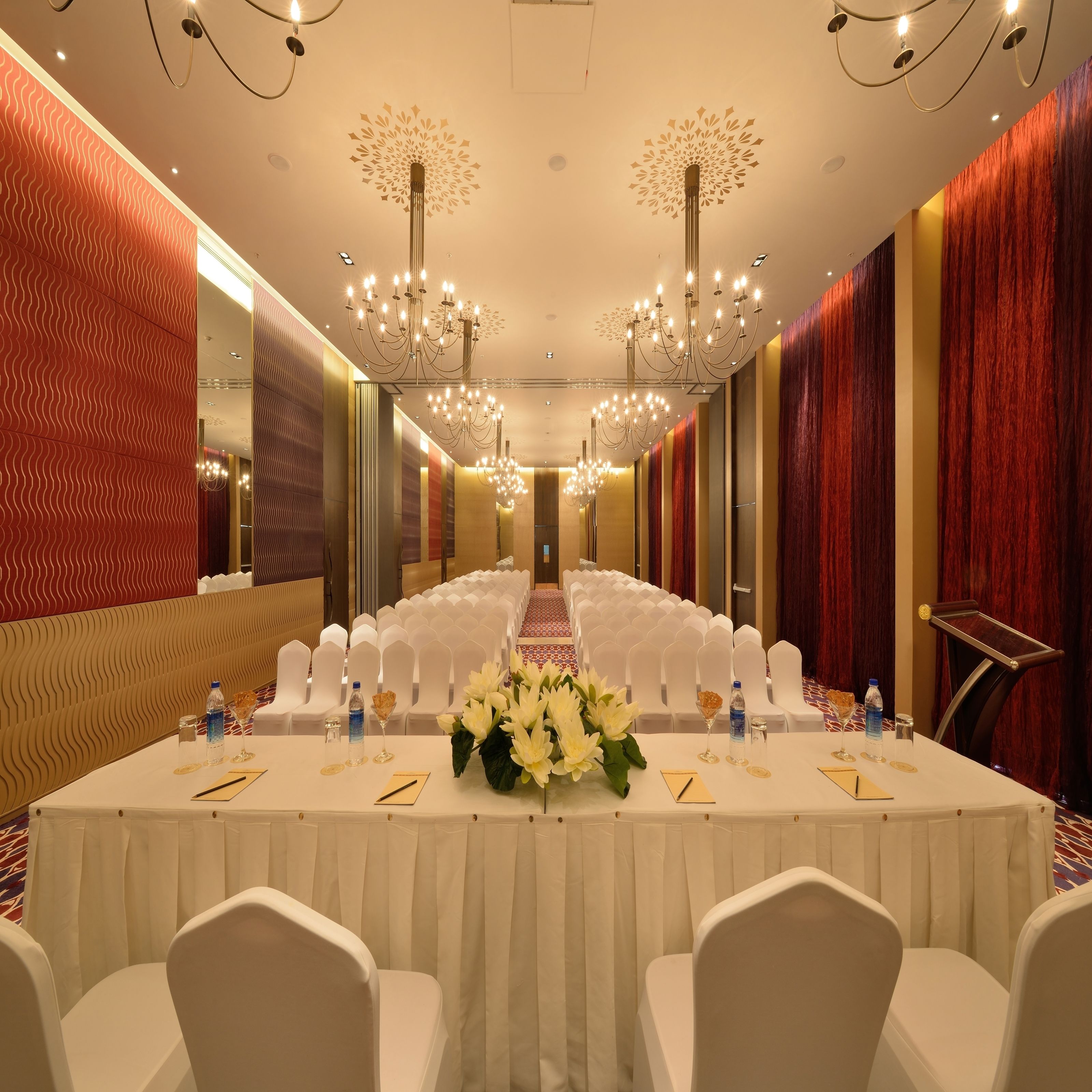 A long banquet hall with rows of white chairs, a central table with floral arrangements, and a chandelier.
