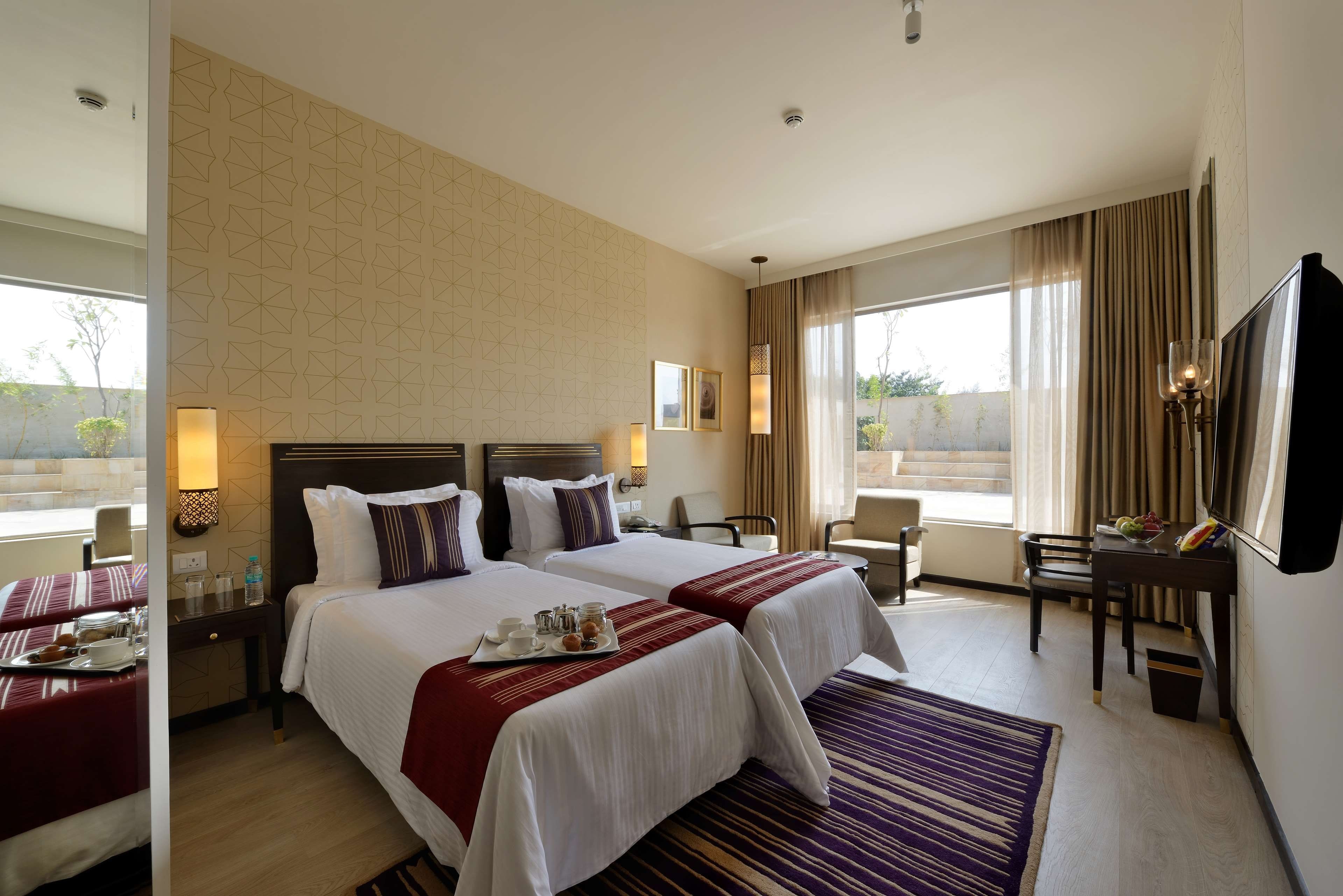 A hotel room with two beds, a desk, a chair, a TV, and a large window with curtains.