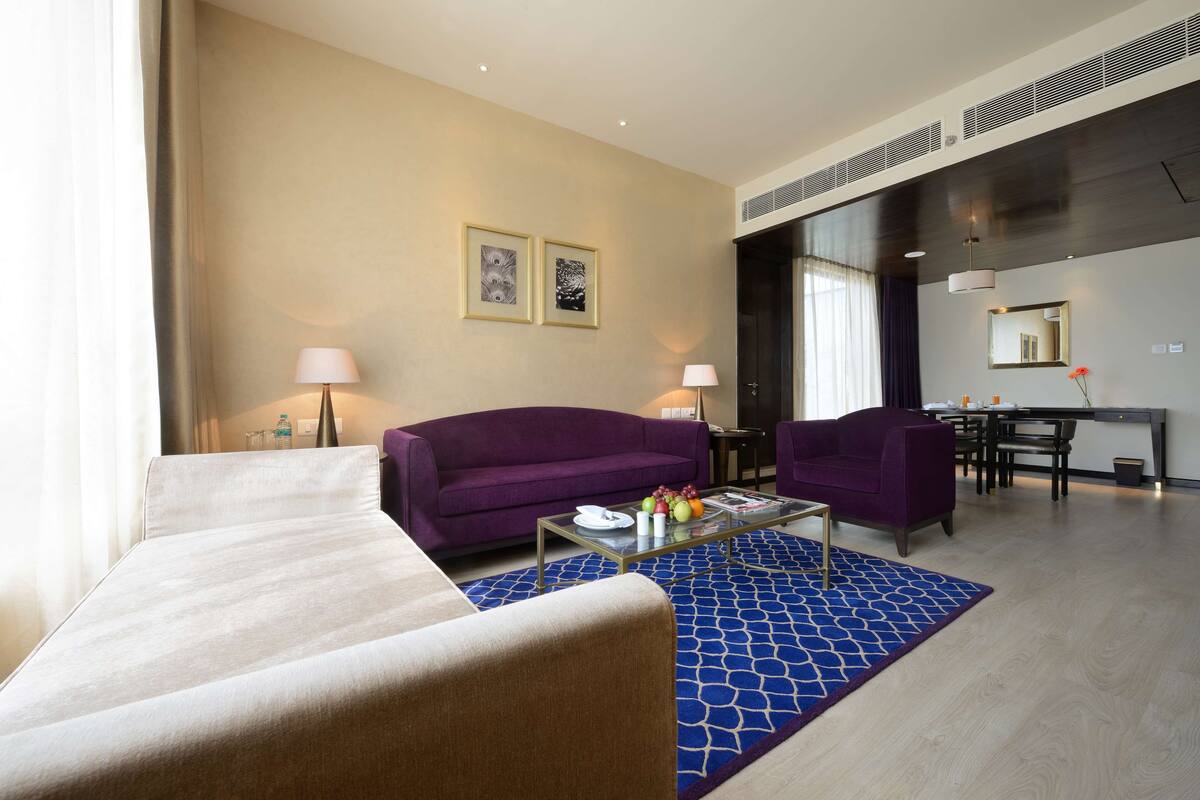 Ett modernt hotellrum med en beige soffa, en lila fåtölj, ett glasbord med frukt och en blå, mönstrad matta.