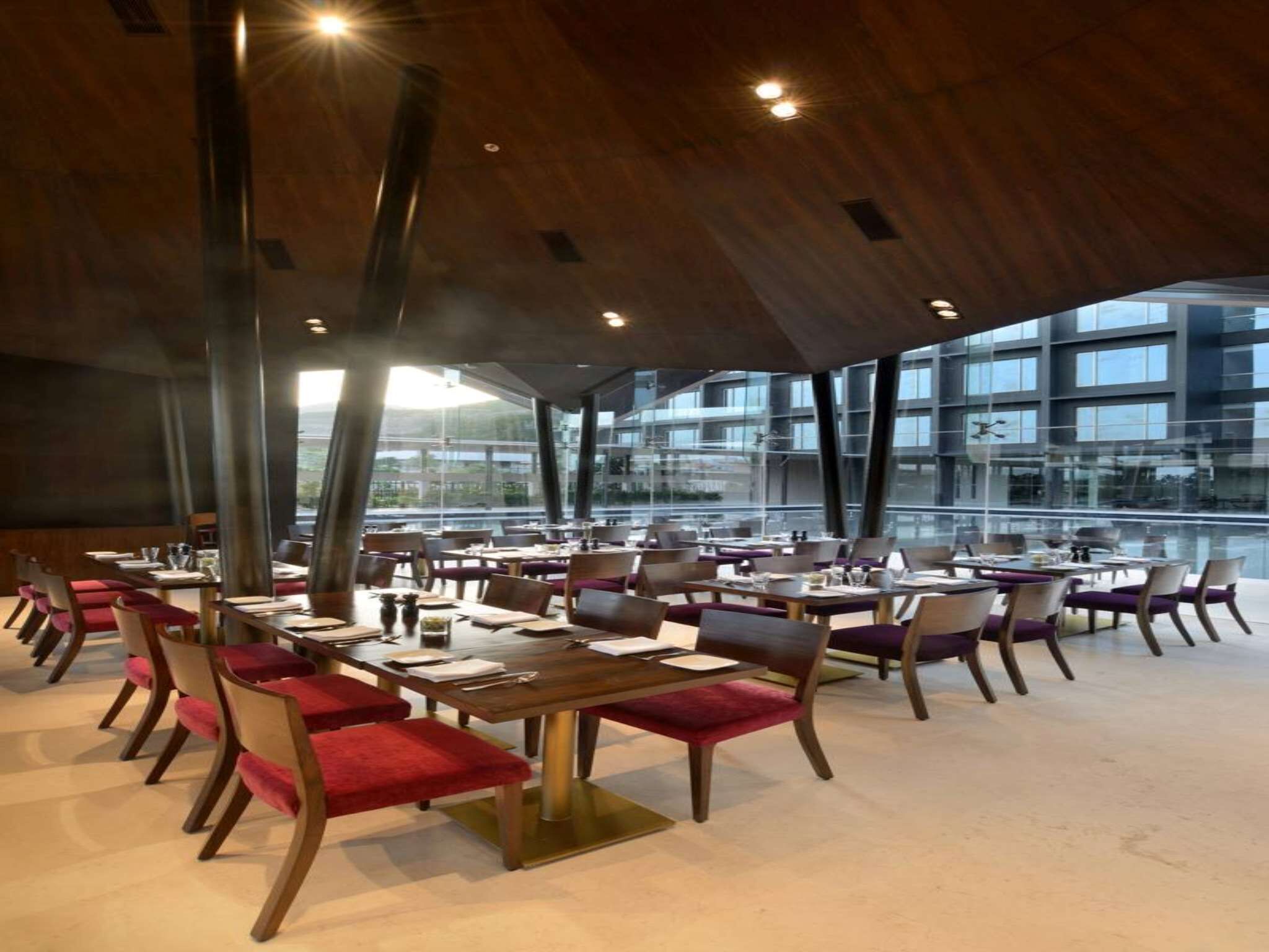 Un restaurante moderno con techo alto y ventanas amplias que ofrecen vistas a un paisaje urbano. Las mesas están dispuestas ordenadamente con sillas tapizadas en rojo.