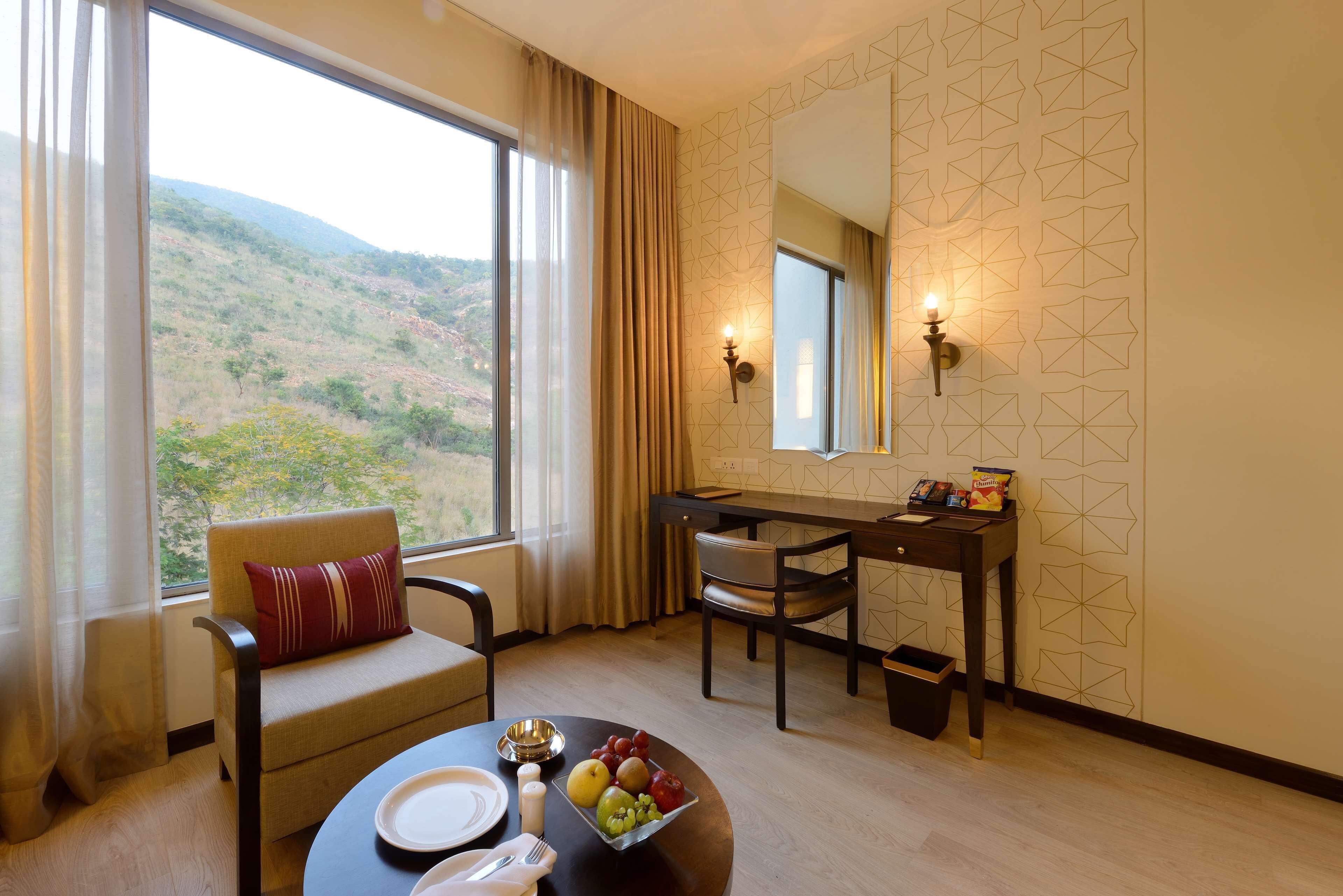 A hotel room with a dining area, a view of the hills, a dining table with plates and fruits, and a chair with a red pillow.