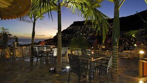 An outdoor seating area with tables and chairs, palm trees, and a view of the ocean at dusk. - Apartamentos Curasol (Mogan)