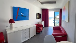 A modern hotel room with a white console, red seating, a TV, and a large window with a view of the sea. - Apartamentos Curasol (Mogan)
