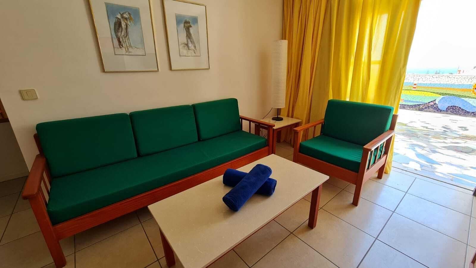 A room with a green sofa, a matching armchair, a small table, and a lamp. There are two framed pictures on the wall.