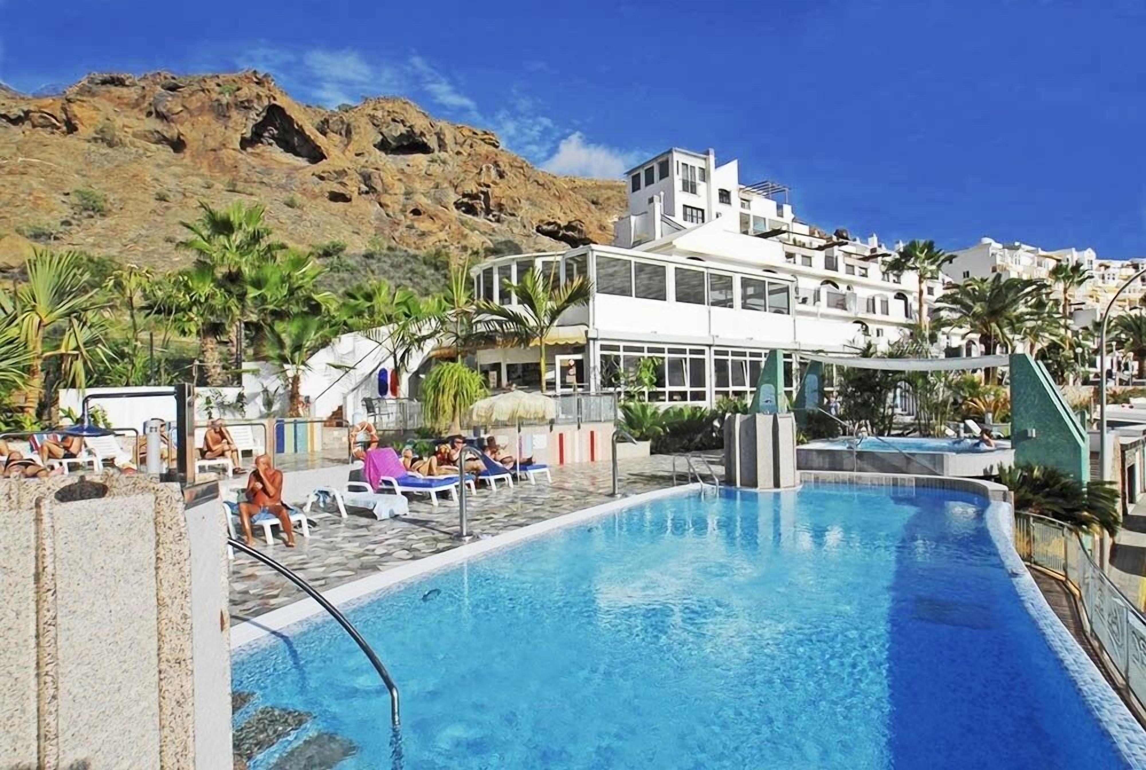 A swimming pool with lounge chairs and palm trees, surrounded by buildings and a rocky hillside.