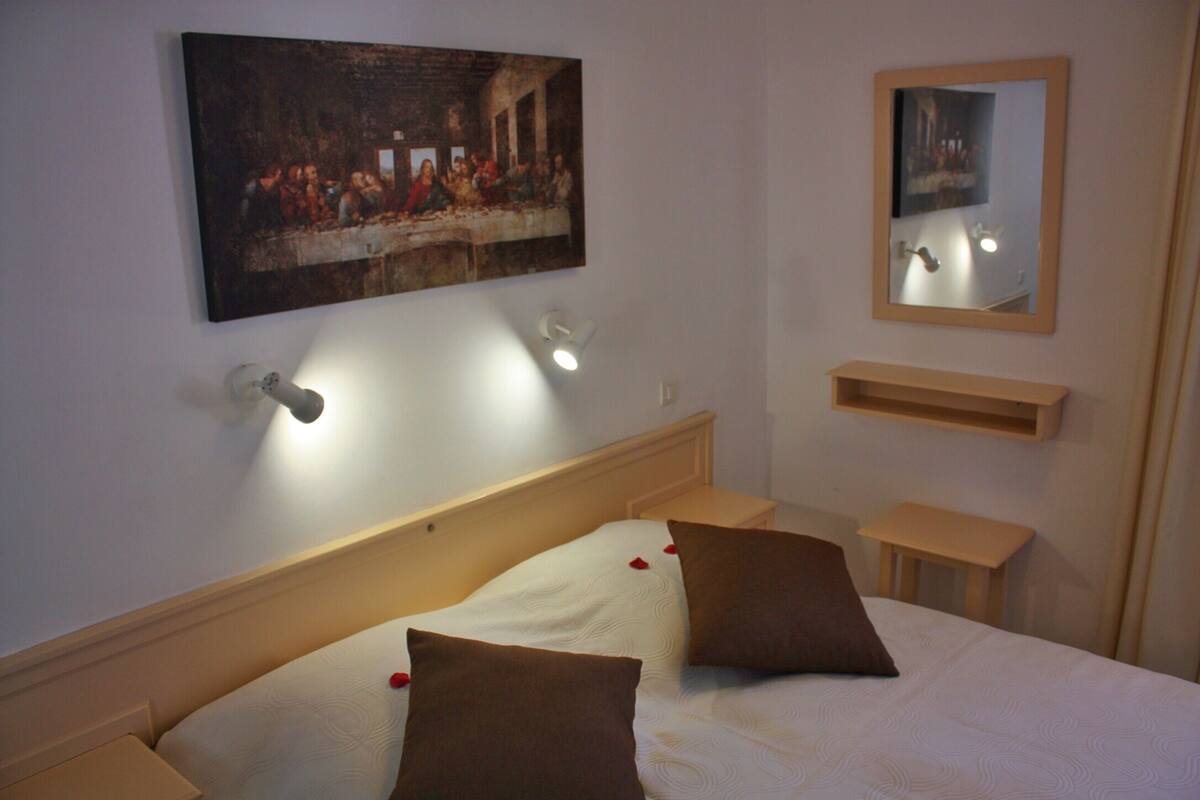 A hotel room with a bed, a painting of 'The Last Supper' on the wall, a mirror, and bedside tables.