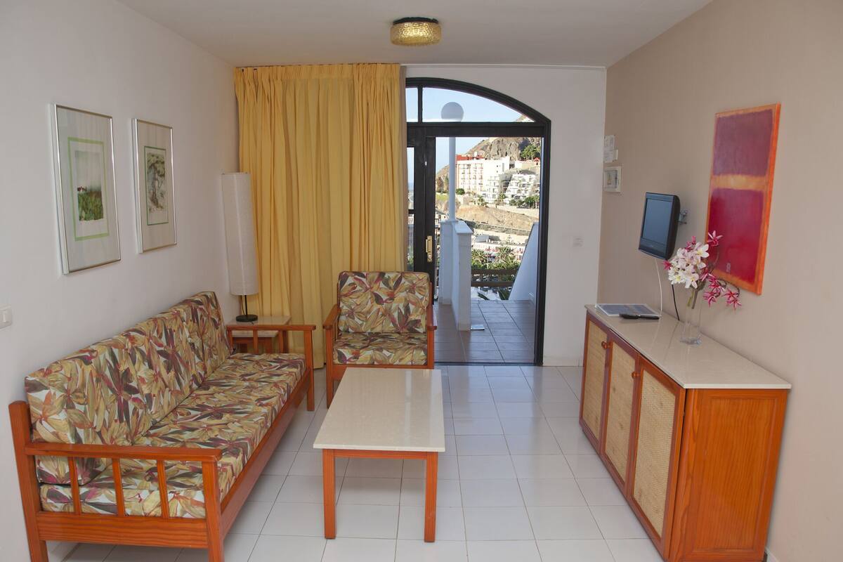 A living room with a sofa, armchair, and coffee table, a TV, and a view of a balcony and buildings.